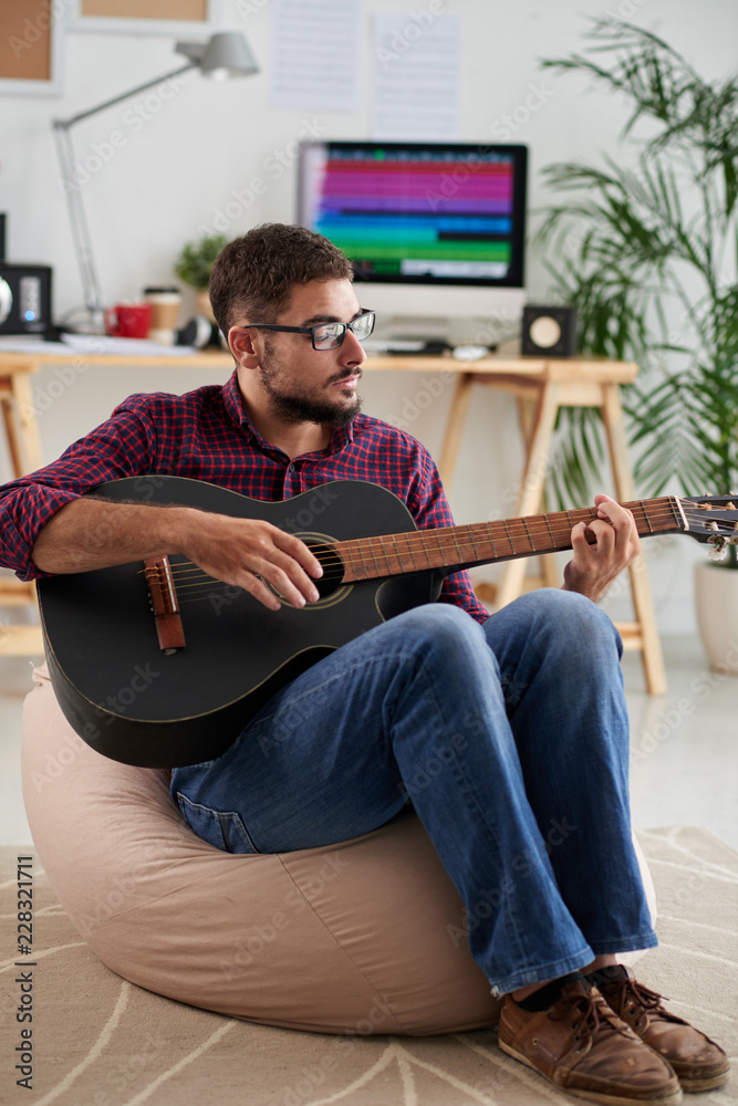 Young man in eyeglasses sitting on sofa and playing guitar