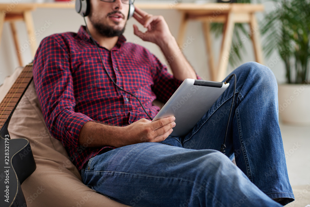 Young man in casual clothing resting on sofa with tablet pc, he wearing headphones and watching something on touchpad