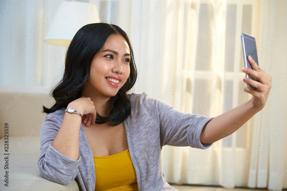 Charming Filipino lady smiling and using smartphone to take selfie while sitting near comfortable sofa in cozy living room Charming Filipino lady smiling and using smartphone to take selfie while sitting near comfortable sofa in cozy living room