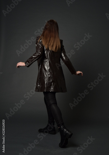 full length portrait of brunette woman wearing long leather coat. standing pose with back to the camera, on grey studio background.