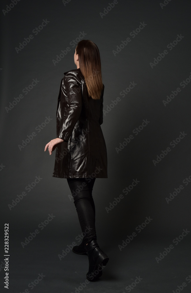 full length portrait of brunette woman wearing long leather coat. standing pose with back to the camera, on grey studio background. full length portrait of brunette woman wearing long leather coat. standing pose with back to the camera, on grey studio background.