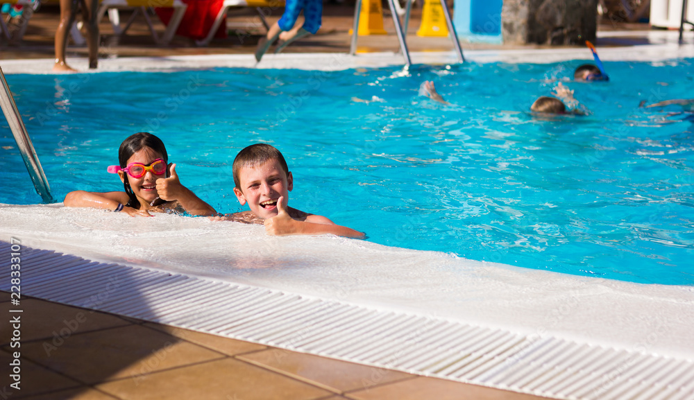 Little boy and girl with pink goggles smiling with thumbs up by swimming pool edge. Young kids having fun on summer holidays. Resort leisure time, travel vacation, hotel activity, tourism concepts
