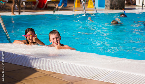 Little boy and girl with pink goggles smiling with thumbs up by swimming pool edge. Young kids having fun on summer holidays. Resort leisure time, travel vacation, hotel activity, tourism concepts