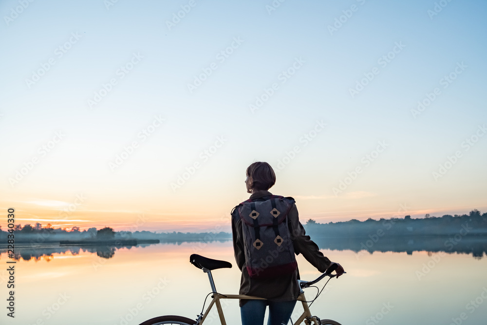 Female cyclist enjoying beautiful blue hour scene by the lake. Woman stands with bike and looks at beautiful lake and sunset