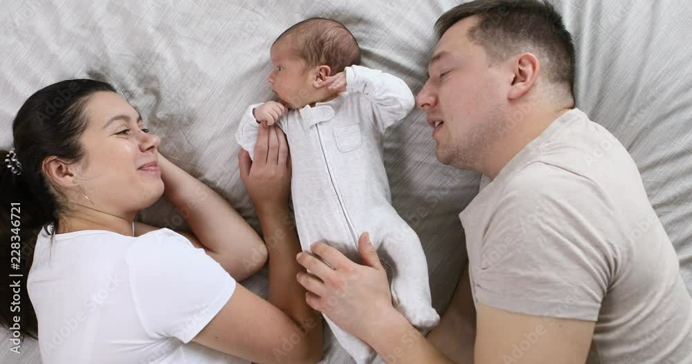 Happy young caucasian parents with their newborn baby lying on the bed at the infant first days at home, top view. Parenthood, love and tender concept Happy young caucasian parents with their newborn baby lying on the bed at the infant first days at home, top view. Parenthood, love and tender concept