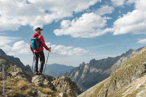 Photography A bearded man in sunglasses and a cap with a backpack stands on top of a rock and looks into a rocky valley high in the mountains
