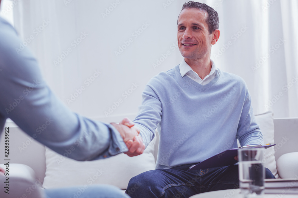 Obraz premium Successful appointment. Waist up of optimistic skilled psychologist holding a clipboard with necessary information while shaking hands with his loyal client