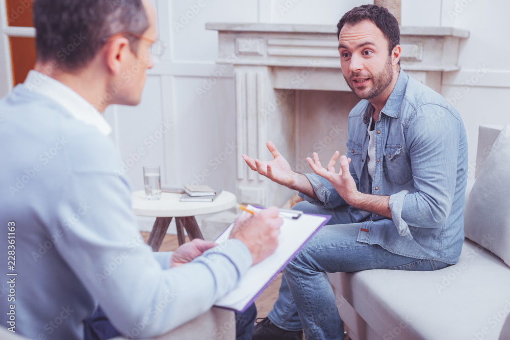 Obraz premium Much information to tell. Excited enthusiastic bearded man explaining his personal problems to psychologist while sitting on sofa and gesticulating