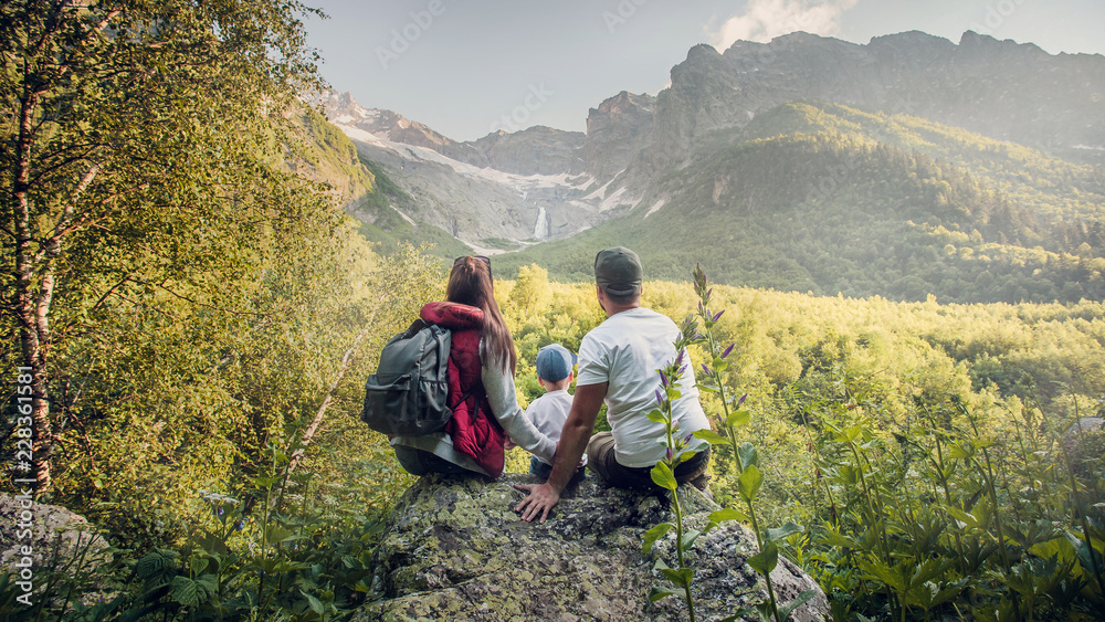 A young European family of three, mom, dad and two-year-old son sit on a rock and look at the mountain waterfall. the view from the back. Russia, Caucasus, Dombay