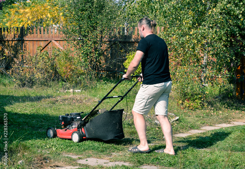 Gardener with electric lawn mower trimming lawn. Seasonal hard work. Summer, autumn, sunny day. Suburb, village, country place. Adult man pruning and landscaping garden, trimming grass, lawn, paths.