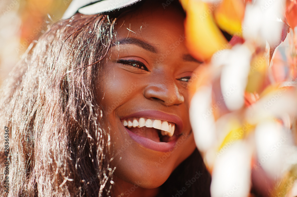 Fototapeta premium Stylowa Afroamerykanin dziewczyna w czapce pozowała w słoneczny jesienny dzień na czerwonych liściach Kobieta model Afryki.