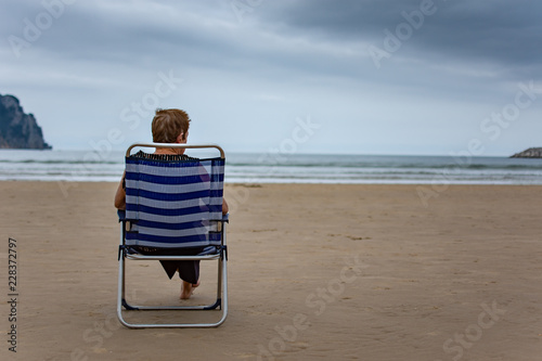 Lonely Elderly woman sitting in a chair, contemplatively gazing at the sea on a cloudy day. She is seen from behind, with sand, the sea, and clouds all around. photo