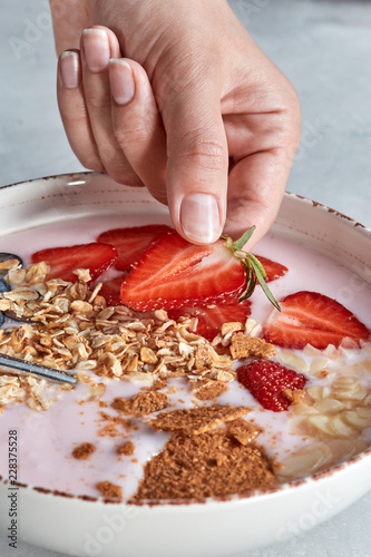 Womans hand puts slices of strawberry to a bowl with natural organic food - smoothie of fruits, nuts, muesli on a gray background. photo