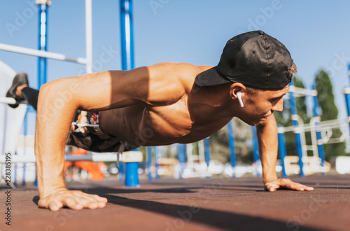 Closeup horizontal image of young athletic muscular man doing push-ups on sports ground outdoor. Caucasian shirtless fitness male doing outdoor workout exercises outside. People and sport concept