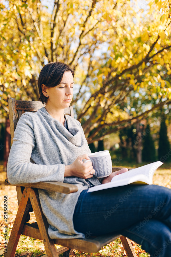 Fototapeta premium Woman wearing warm knit clothes drinking a cup of hot tea or coffee and reading a book outdoors in sunlight - season, literature, leisure time and people concept - beautiful morning in the garden