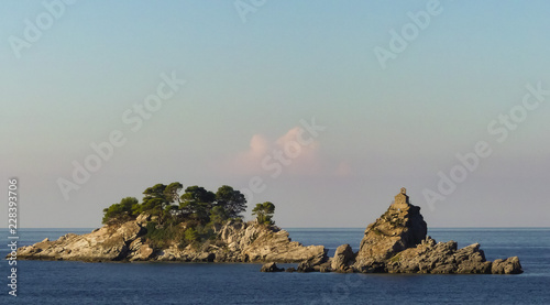 Sunlit view of the islands Katic and Sveta Nedjelja (Holy Sunday) on the bay from Petrovac, Montenegro. Small chapel perched on top of rocks. Small white cloud in background passing between. Blue sky. photo