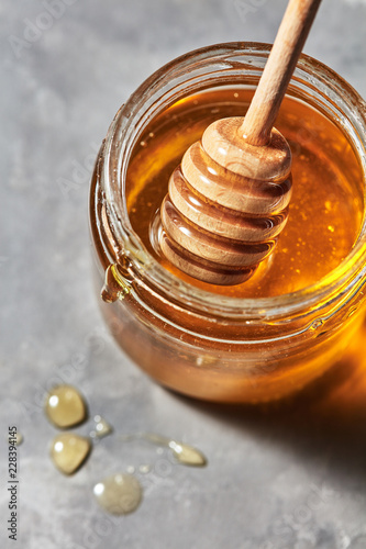 Close up of wooden honey stick in a glass pot with sweet organic dessert on a gray stone background, traditional useful sweetness.