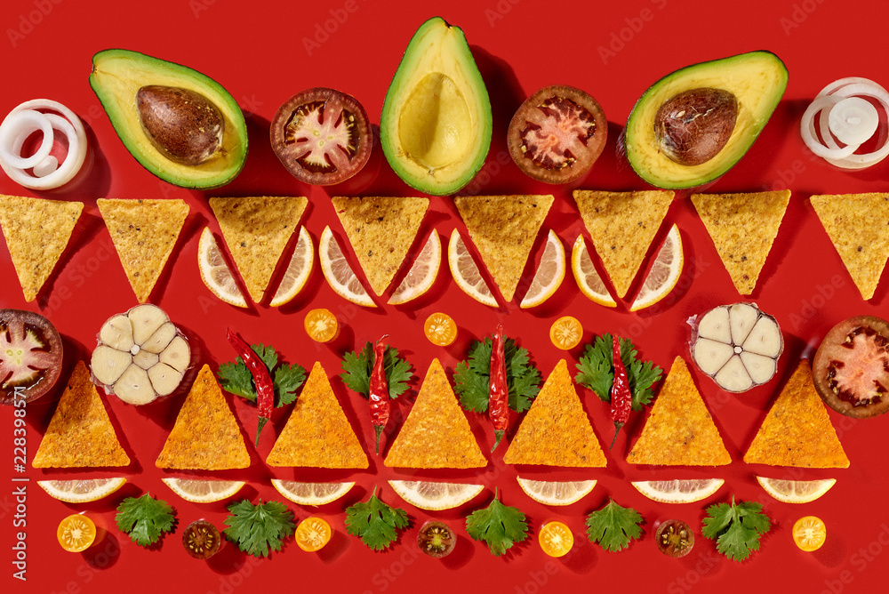 Creative geometric food pattern from mexican nachos corn chips, fresh vegetables, fruits, greens, chili, garlic - ingredients for tomato chili sauce on a red background. Flat lay