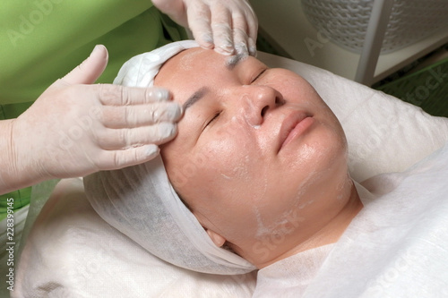 A professional beautician in a green dress washes the face of a relaxed Muslim woman with soap. Eastern girl on the procedure for deep cleaning of the skin. Aesthetic appearance in the beauty salon.