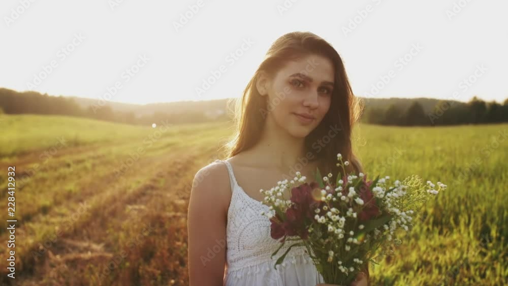 Pretty young woman in white dress standing in a meadow with bouquet of wild flowers on summer evening. Long hair flowing in wind in sunshine at sunset, she looks up into the camera and smiles Pretty young woman in white dress standing in a meadow with bouquet of wild flowers on summer evening. Long hair flowing in wind in sunshine at sunset, she looks up into the camera and smiles