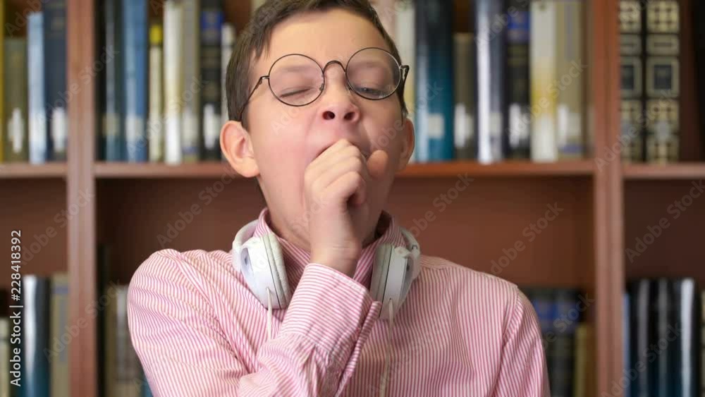portrait shot of the cute yawns schoolboy standing near the bookshelf in the library portrait shot of the cute yawns schoolboy standing near the bookshelf in the library