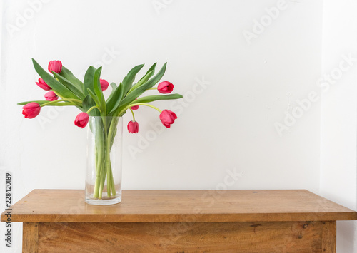 Red tulips with green leaves in glass vase on oak sidetable against white wall with copy space (selective focus)