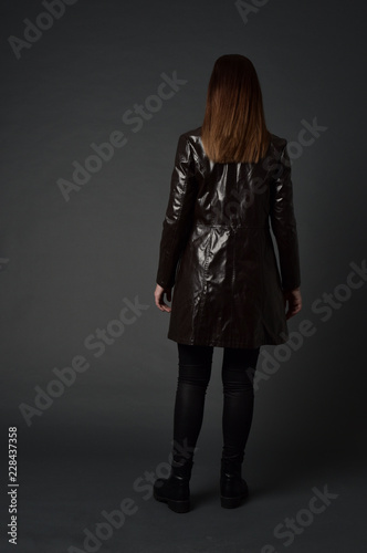 full length portrait of brunette woman wearing long leather coat. standing pose with back to the camera, on grey studio background.