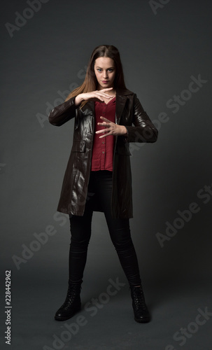 full length portrait of brunette girl wearing long leather coat and boots. standing pose  on grey studio background.