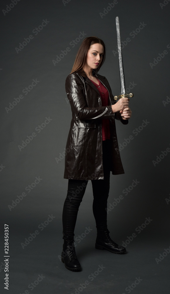 full length portrait of brunette girl wearing long leather coat and boots. standing pose and holding a sword on grey studio background. full length portrait of brunette girl wearing long leather coat and boots. standing pose and holding a sword on grey studio background.