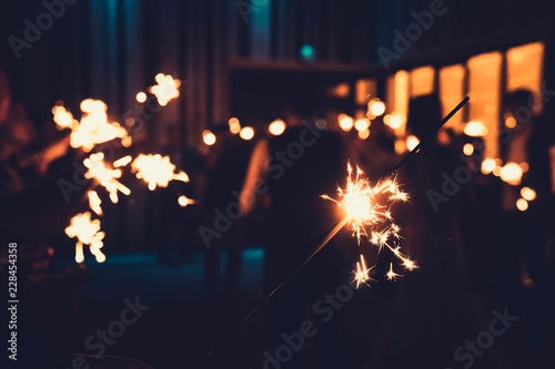 Close up of a bengal light at the blurred background of people standing with burning sparklers in semidarkness. Indoor firework by the turned off light. Indoor celebration with bengal lights burning.