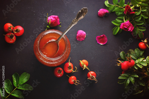 Spoon in a jar of rosehip jam and its berries with leaves nearby, top view