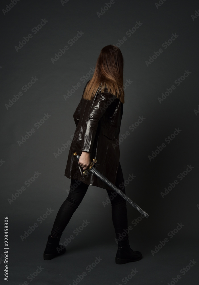 full length portrait of brunette girl wearing long leather coat and boots. standing pose and holding a sword on grey studio background. full length portrait of brunette girl wearing long leather coat and boots. standing pose and holding a sword on grey studio background.