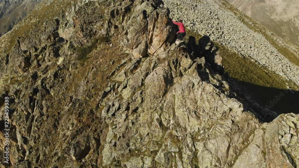 A young guy with a beard, a mountaineer in a cap and sunglasses, climbs a rocky ridge high in the mountains. Parkour in the mountains. Arial view A young guy with a beard, a mountaineer in a cap and sunglasses, climbs a rocky ridge high in the mountains. Parkour in the mountains. Arial view