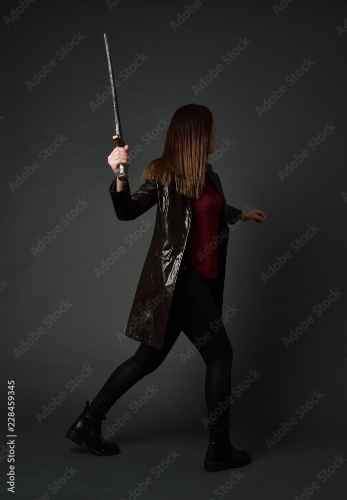 full length portrait of brunette girl wearing long leather coat and boots. standing pose and holding a sword on grey studio background. full length portrait of brunette girl wearing long leather coat and boots. standing pose and holding a sword on grey studio background.