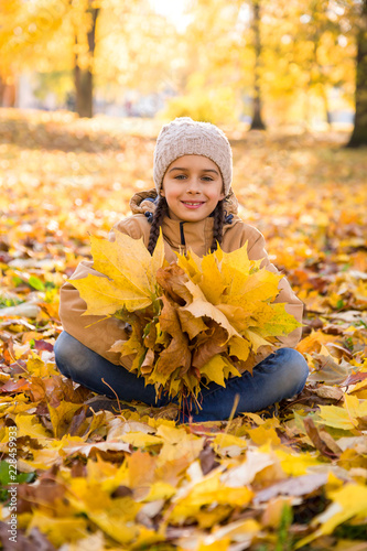 Autumn portrait  Cute smiling little girl sitting on the fallen leaves in autumn park and holding the bouquet of yellow maple leaves in her hands  vertical 