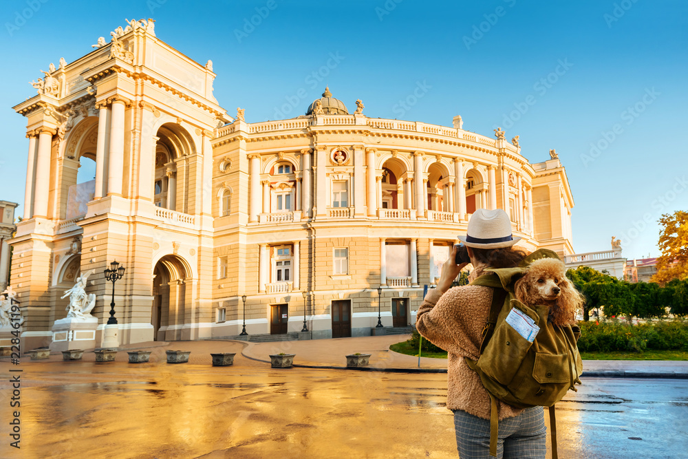 Woman traveler with dog in the backpack taking pictures with a camera while traveling. Odessa Opera Theatre.	