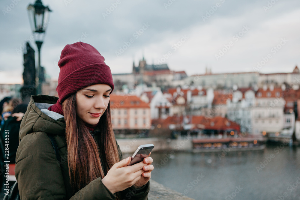 A young beautiful tourist girl stands on the Charles Bridge in Prague in the Czech Republic and uses a mobile phone to call or view a map or mobile application.