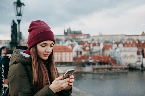 Obraz na płótnie A young beautiful tourist girl stands on the Charles Bridge in Prague in the Czech Republic and uses a mobile phone to call or view a map or mobile application. w salonie