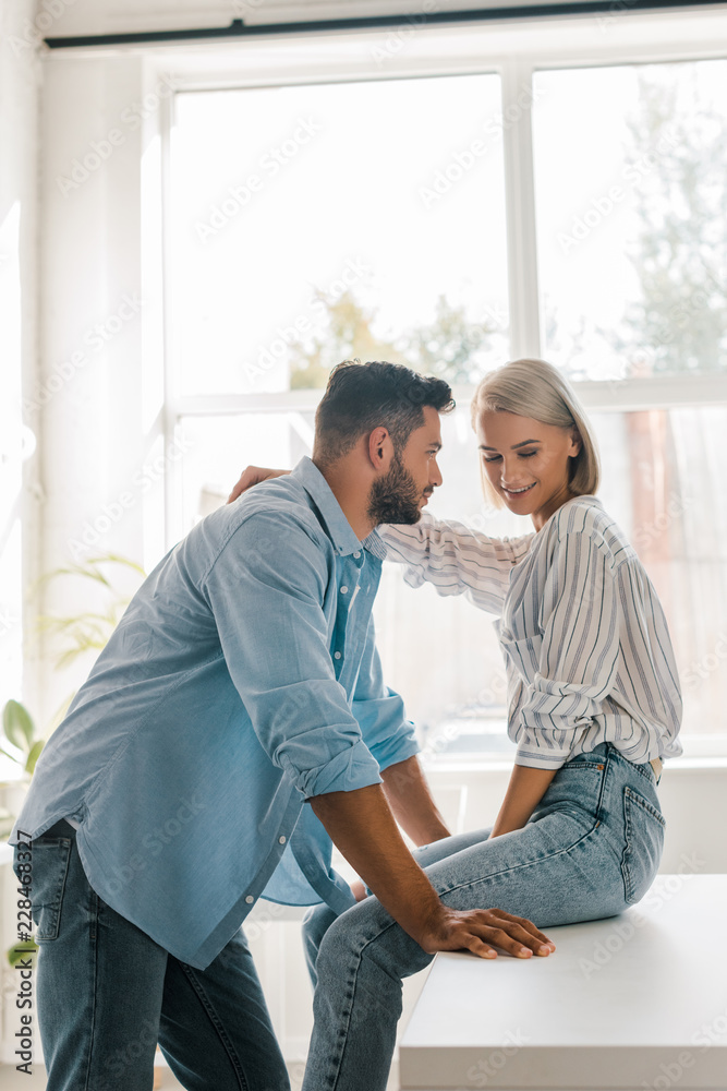 side view of smiling young couple hugging in kitchen while girlfriend sitting on kitchen counter side view of smiling young couple hugging in kitchen while girlfriend sitting on kitchen counter