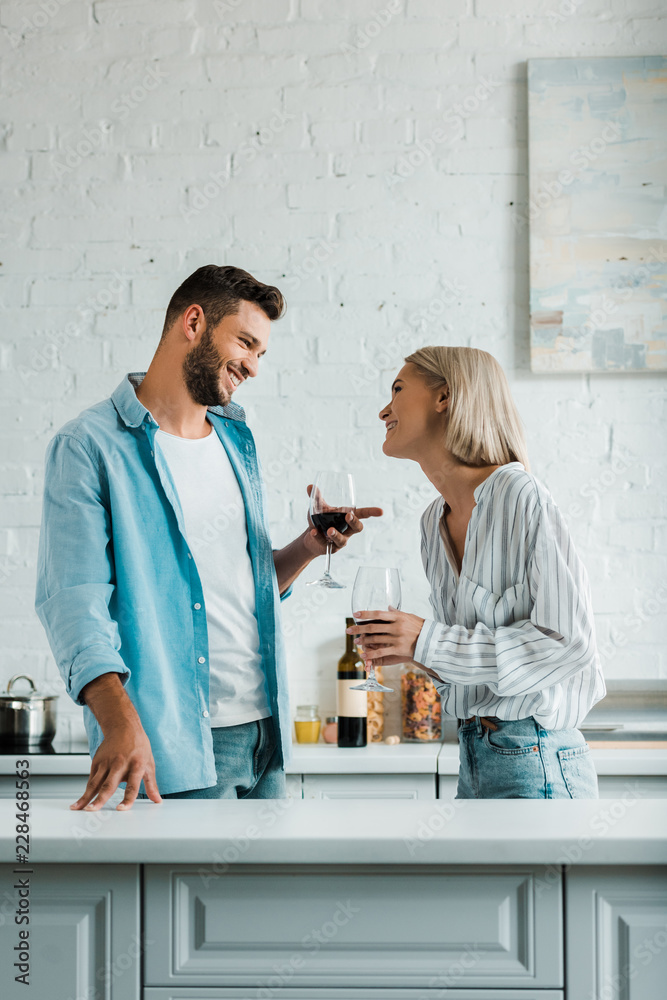 Fototapeta premium smiling young couple talking, holding glasses of red wine and looking at each other in kitchen
