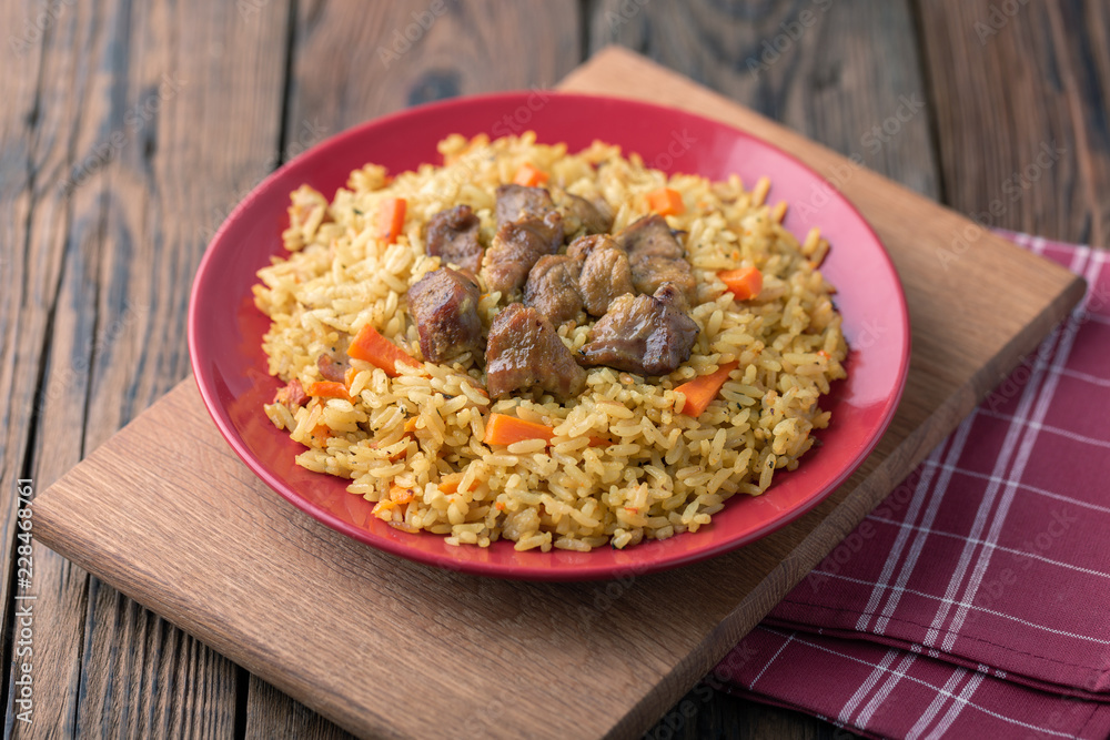 Red plate with pilaf on a brown wooden table. On the table is red pepper, green onions, garlic, cherry tomatoes, red napkin, spoon. Red plate with pilaf on a brown wooden table. On the table is red pepper, green onions, garlic, cherry tomatoes, red napkin, spoon.