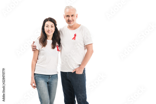 smiling adult interracial couple in blank t-shirts with aids awareness red ribbons embracing and looking at camera isolated on white