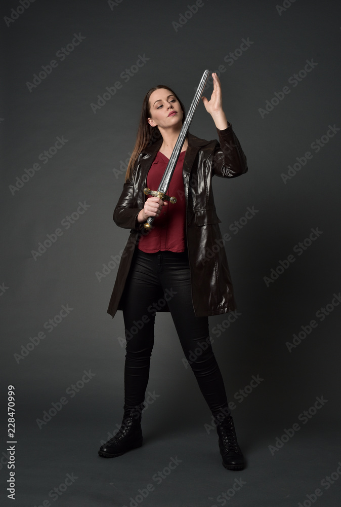 full length portrait of brunette girl wearing long leather coat and boots. standing pose and holding a sword on grey studio background. full length portrait of brunette girl wearing long leather coat and boots. standing pose and holding a sword on grey studio background.
