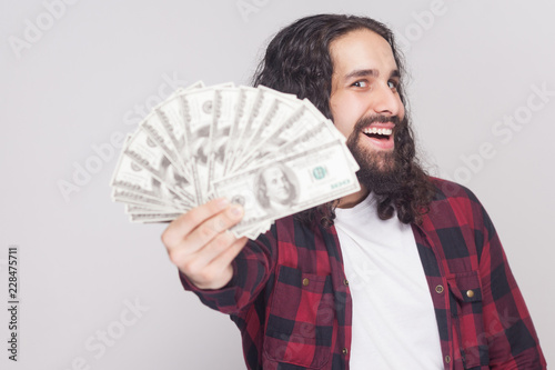 Take it! Happy kidness young businessman in red checkered shirt and long curly hair standing, holding many dollars looking at camera with open mouth. Indoor studio shot, isolated on gray background photo