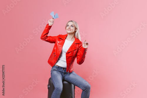 Happy young woman with travel suitcase launches paper airplane. Blonde tourist girl on pink background, concept
