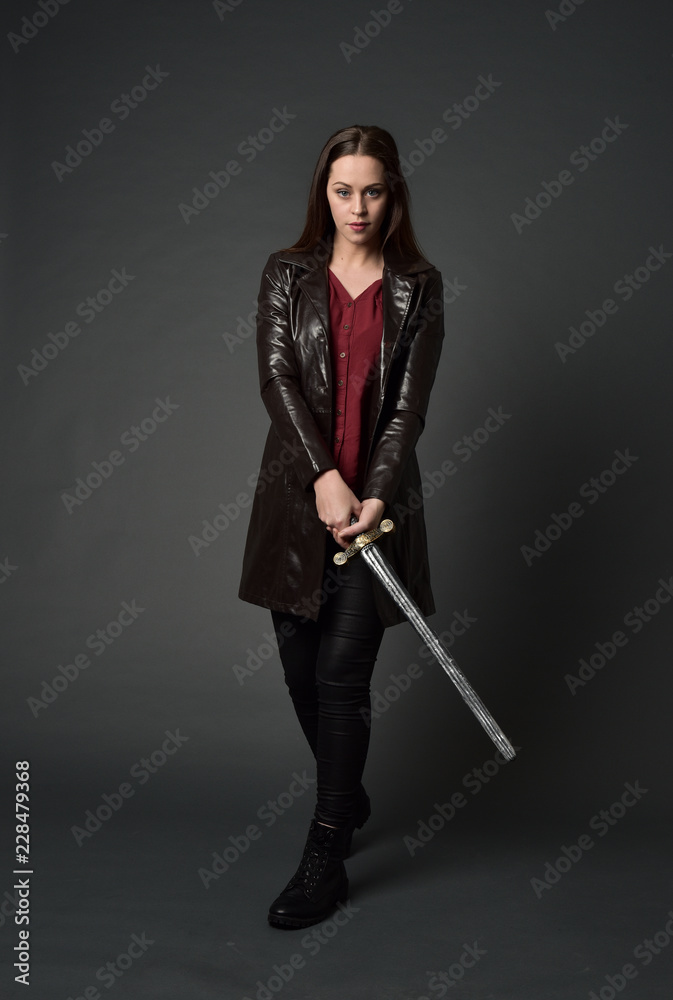 full length portrait of brunette girl wearing long leather coat and boots. standing pose and holding a sword on grey studio background.