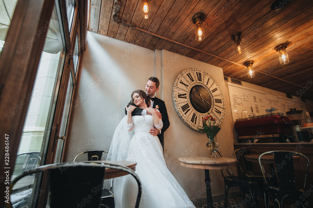 Beautiful newlyweds stand on the background of a big clock and look out the window. The newlyweds embrace in an old cafe with lamps.