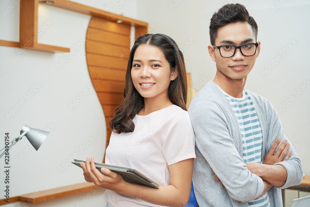Obraz premium Trendy modern Asian man and woman with tablet standing back to back in office looking confidently at camera