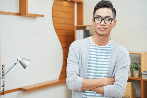Casual young Asian man in black glasses standing with arms crossed looking at camera in office