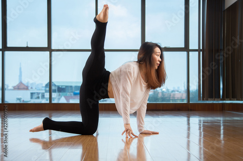 Side view of beautiful Asian female lifting leg while performing contemporary dance moves on studio floor near window